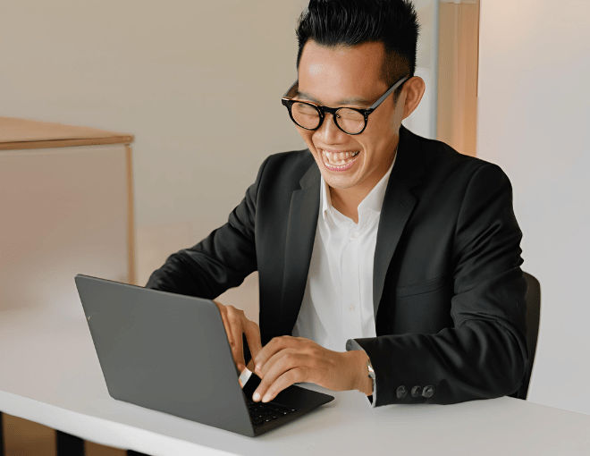Wearing a crisp white shirt and black blazer, an Indonesian man types on his laptop, his face reflecting enjoyment and satisfaction.